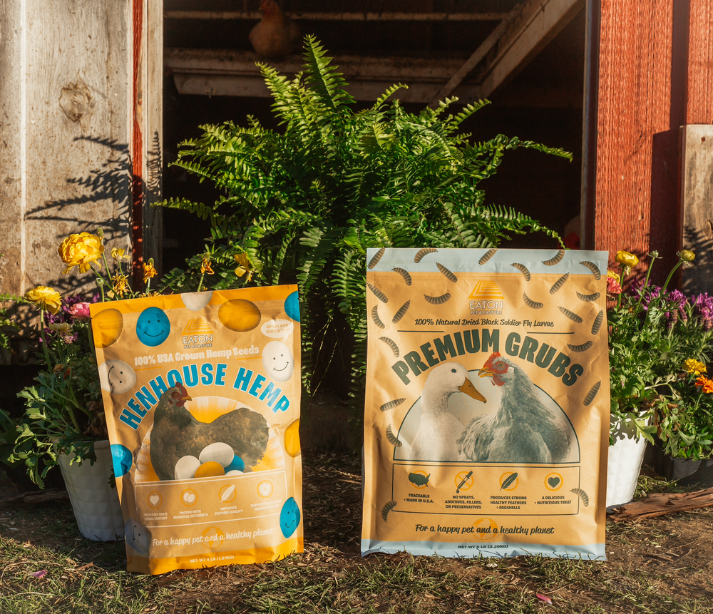 Two 9lb treat bags for chickens: Grubs and Hemp by Eaton Pet and Pasture, both with colorful packaging against a garden back