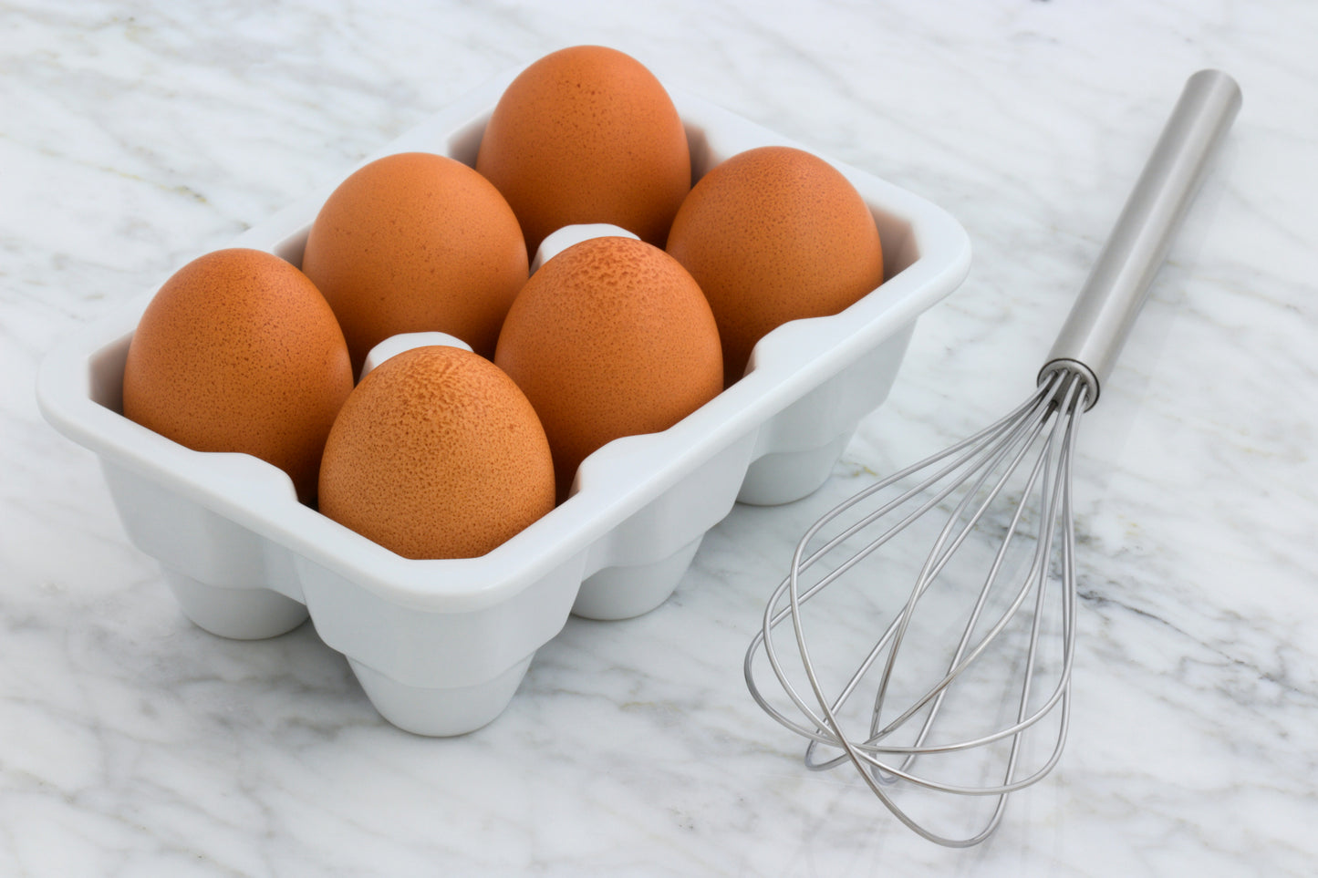 Brown eggs in a white carton with a stainless steel whisk beside them